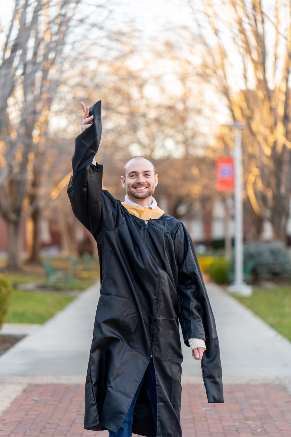 Graduate in a black gown raising his arm on a tree-lined campus