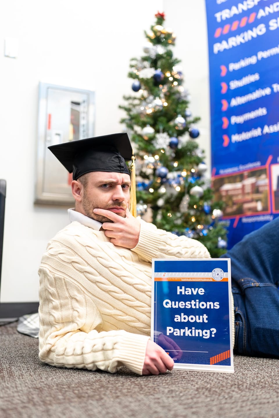 Graduate sitting by a Christmas tree holding a humorous sign