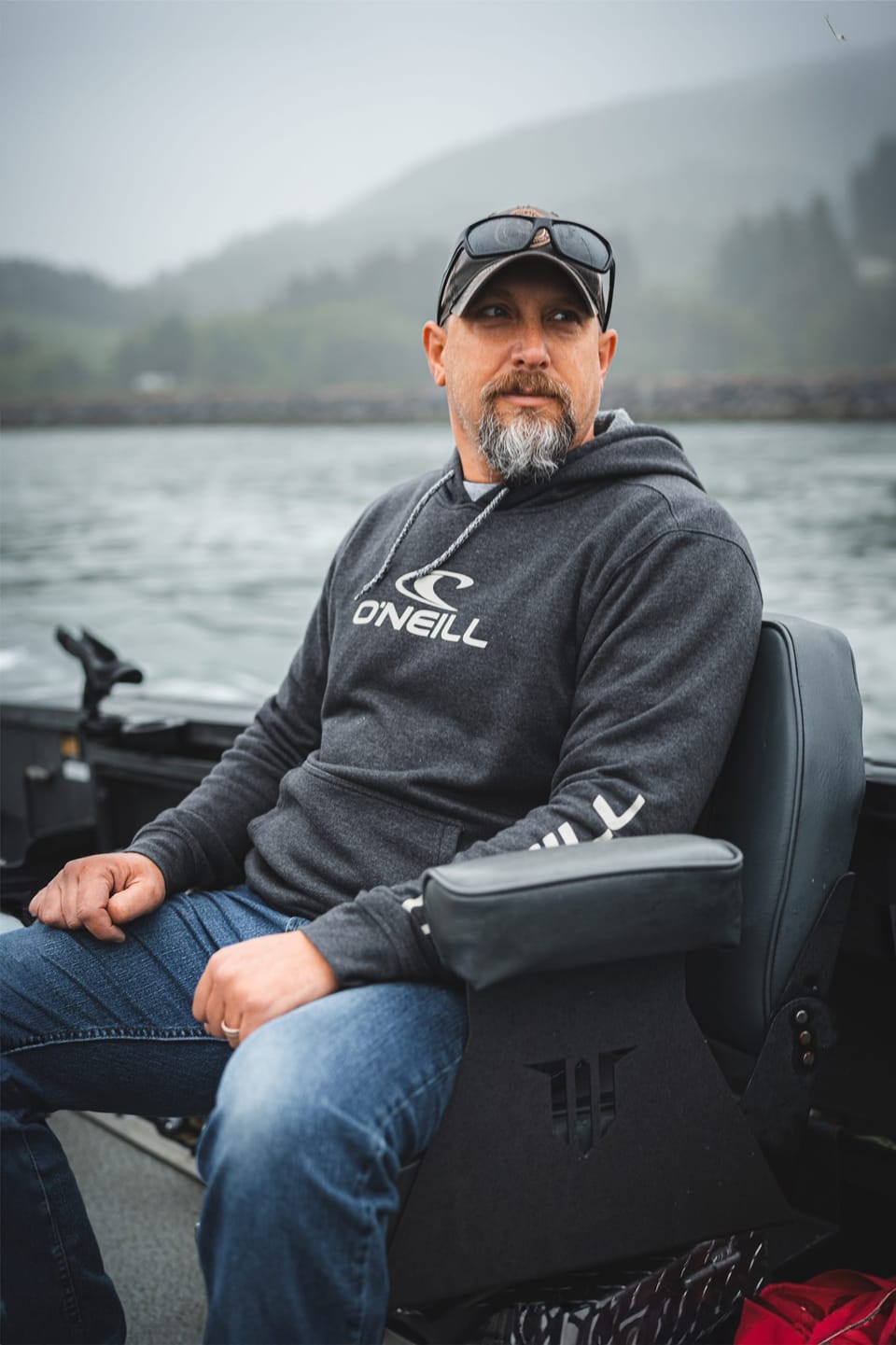 Man in a gray hoodie seated in a boat with mountains behind