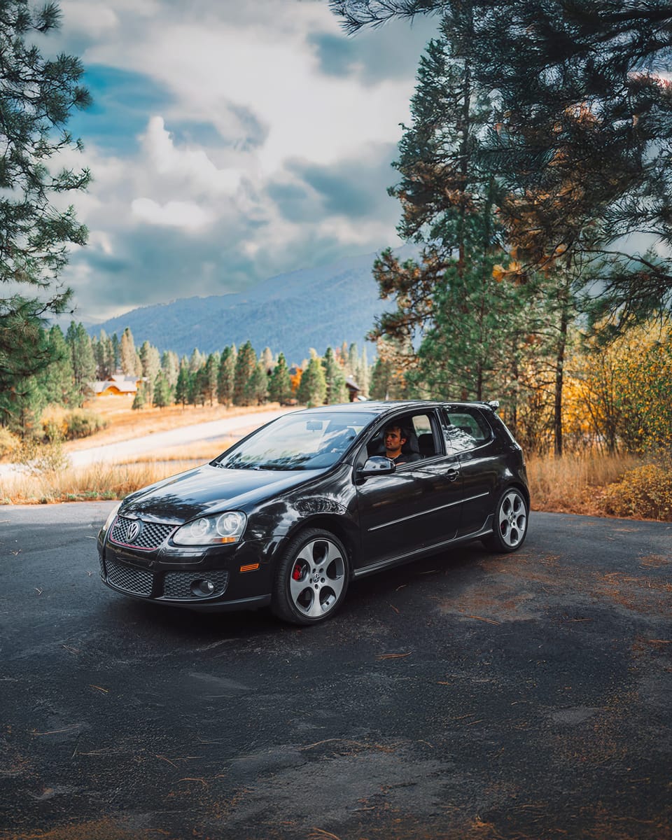 Driver leaning out of a black VW GTI Mk5 among pines and fall-colored trees below a mountain